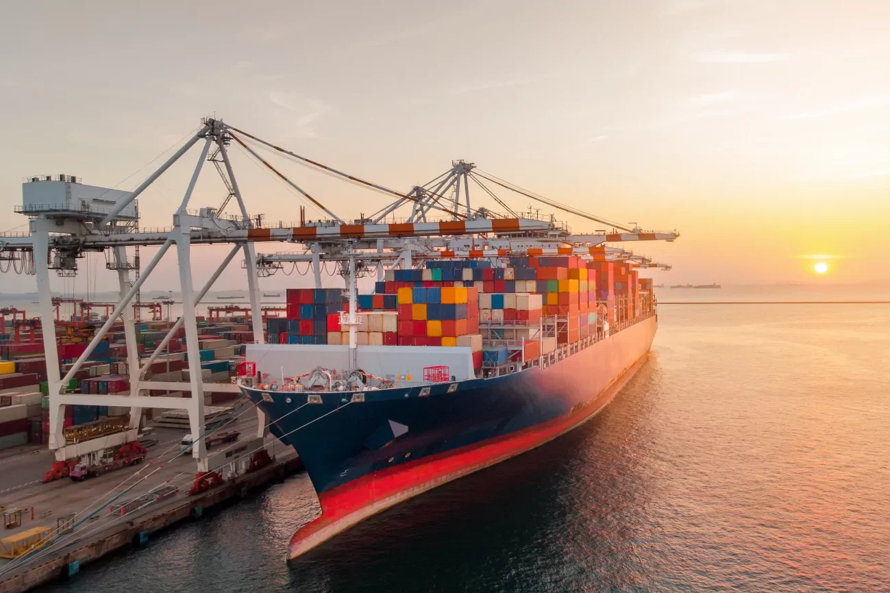 Mit bunten Containern beladenes Frachtschiff liegt im Hafen, im Hintergrund geht die Sonne unter.